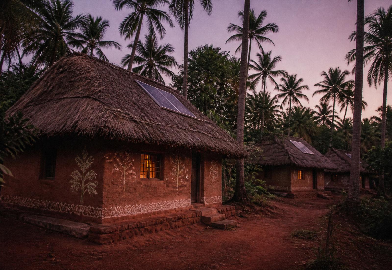 Earthen cob houses with thatched roofs and small solar panels at dusk